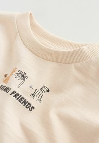 T-shirt en coton beige clair avec encolure côtelée, broderie d'animaux de safari et texte "SAFARI FRIENDS" en noir.