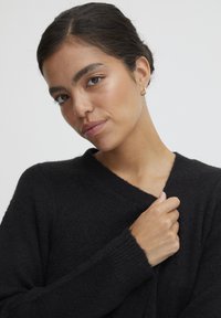 Schwarzer, strukturierter Pullover mit V-Ausschnitt und langen Ärmeln, lässig getragen. Die Hand des Models ruht auf dem Stoff und hebt die weiche Textur hervor.