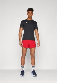Schwarzes Sport-T-Shirt und rote Shorts mit weißen Akzenten. Das Modell trägt dunkle Sneakers mit weiß-schwarz gestreiften Socken. Einfaches Design.