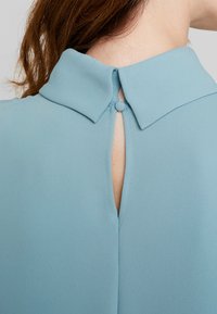 Ljusblå blus med krage, med knapppåsättning och en nyckelhålsöppning bak. Slät, flödande tyg med en subtil lyster.