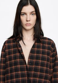 Camisa de cuadros negra y naranja, de manga larga con cuello, que presenta cierre de botones y un bolsillo frontal. Tejido ligero con un aspecto texturizado.