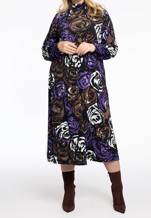 Vrouw draagt een lange mouw, hoog gesloten jurk met paarse, bruine en witte abstracte bloemmotieven, gecombineerd met bruine enkellaarsjes.