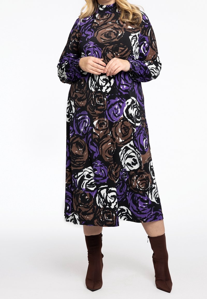 Femme portant une robe à manches longues et col haut avec des motifs floraux abstraits en violet, marron et blanc, associée à des bottines marron.