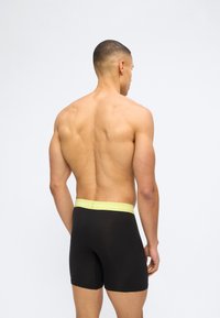 Μυώδης άνδρας με κοντό κούρεμα που φοράει μαύρα μπόξερ με ζώνη μέσης της Calvin Klein σε κίτρινο χρώμα, στέκεται μπροστά σε απλό φόντο.