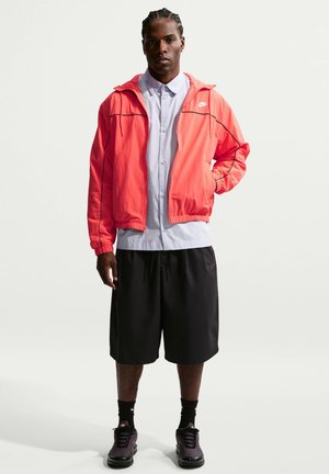 Uomo con trecce che indossa un giubbotto antivento Nike corallo acceso, una camicia chiara con bottoni, pantaloncini neri, calzini neri e scarpe da ginnastica scure, in piedi su sfondo bianco.