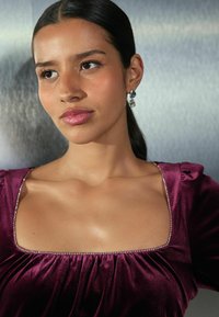 Haut en velours violet avec un décolleté carré, orné de tissu froncé et d'accents en bijoux le long du décolleté. Peau lisse et cheveux longs.