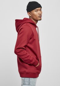 Rød hettegenser med kengurulomme, ribbede ermer og nederkant; kombinert med lyseblå jeans og en svart lue.