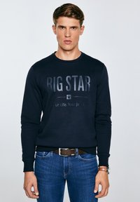 Jaunas vyras dėvintis tamsiai mėlyną „BIG STAR“ džemperį, mėlynas džinsas su rudu diržu ir rankinį laikrodį, stovintis priešais paprastą šviesią fono sieną.