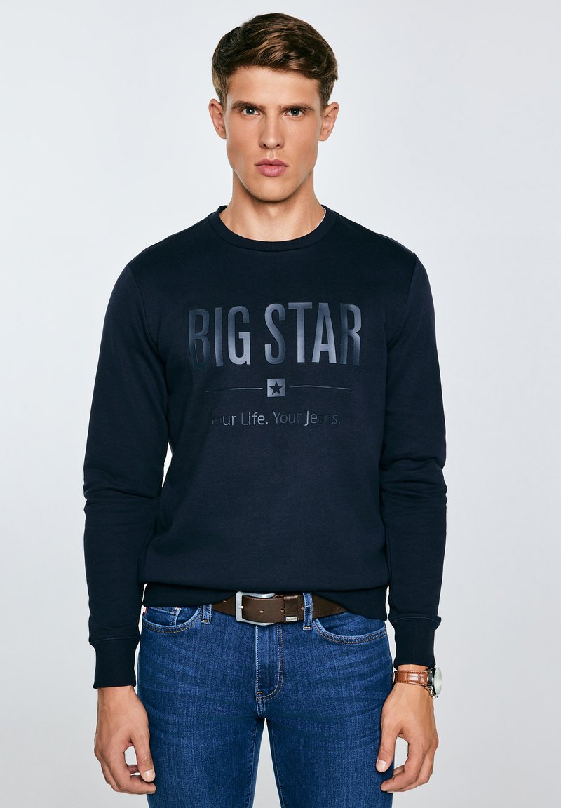 Jaunas vyras dėvintis tamsiai mėlyną „BIG STAR“ džemperį, mėlynas džinsas su rudu diržu ir rankinį laikrodį, stovintis priešais paprastą šviesią fono sieną.