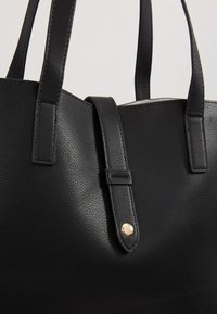 Sac fourre-tout en cuir noir avec poignées doubles et une lanière centrale dotée d'un détail de fermeture à bouton doré.