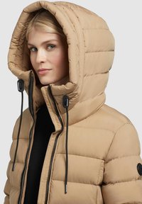 Beige Daunenjacke mit großer, gesteppten Kapuze, Reißverschluss und verstellbaren Kordelzügen. Glatte Textur mit dezentem Schimmer.