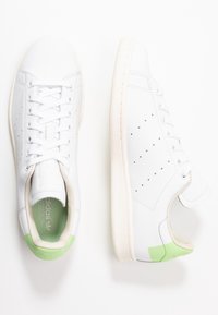 Vita Adidas-sneakers med ljusgröna detaljer på hälen, perforerade sidodetaljer och vita snören, visade från ovan- och sidovy på en vit bakgrund.