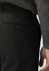 Close-up van zwarte op maat gemaakte broek met een knoopzak aan de achterkant, gedragen met een grijze geribde trui en een zichtbare hand aan de zijkant.