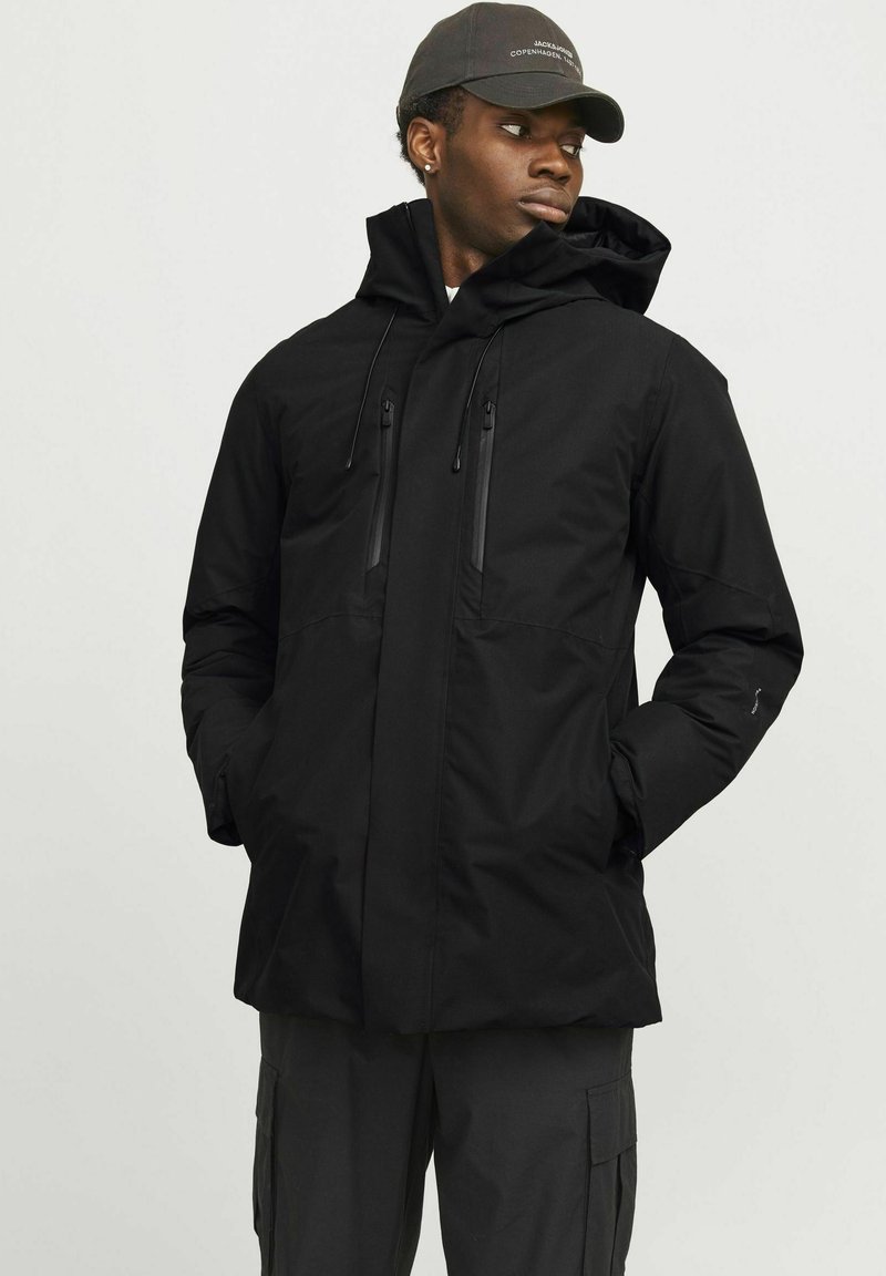 Schwarze, isolierte Jacke mit Kapuze, ausgestattet mit Reißverschlusstaschen auf der Brust und einem eleganten Design. Kombiniert mit dunklen Cargo-Hosen.