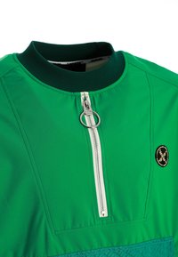 Chaqueta verde con cuello acanalado verde oscuro, cremallera blanca vertical con tirador redondo plateado y un pequeño parche circular negro y amarillo.