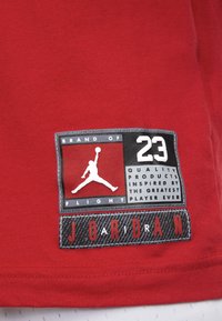 Maglietta rossa in cotone con un'etichetta brandizzata che mostra un logo silhouette bianco, testo nero e uno sfondo testurizzato in bianco e nero.