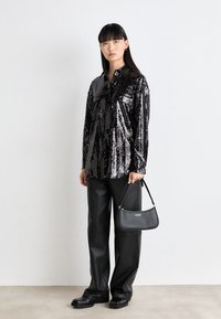 Schwarze Pailletten-Hemdbluse mit langen Ärmeln, kombiniert mit lockeren schwarzen Hosen und einer kleinen schwarzen Handtasche mit Logo. Quadratförmige Schuhe.