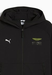 Chaqueta negra con cremallera y capucha, con un logo blanco de Puma y un logo amarillo de Aston Martin. Tela lisa con dos bolsillos frontales.