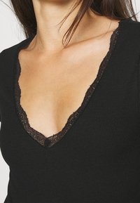 Svart ribbad topp med en djup V-ringning, som har en delikat spetskant längs kanterna. Mjuk textur och figurnära design.