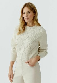 Vrouw met blond haar, gekleed in een crèmekleurige geribbelde trui met diagonale steken en lichtbeige broek, staand met één hand in haar zak.