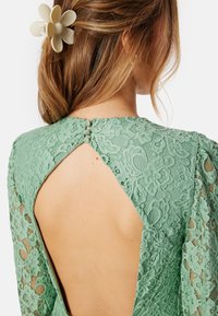 Abito in pizzo verde menta con schiena a chiave, chiusura a bottoni sul collo e fermaglio per capelli floreale, che mette in risalto intricati motivi floreali.