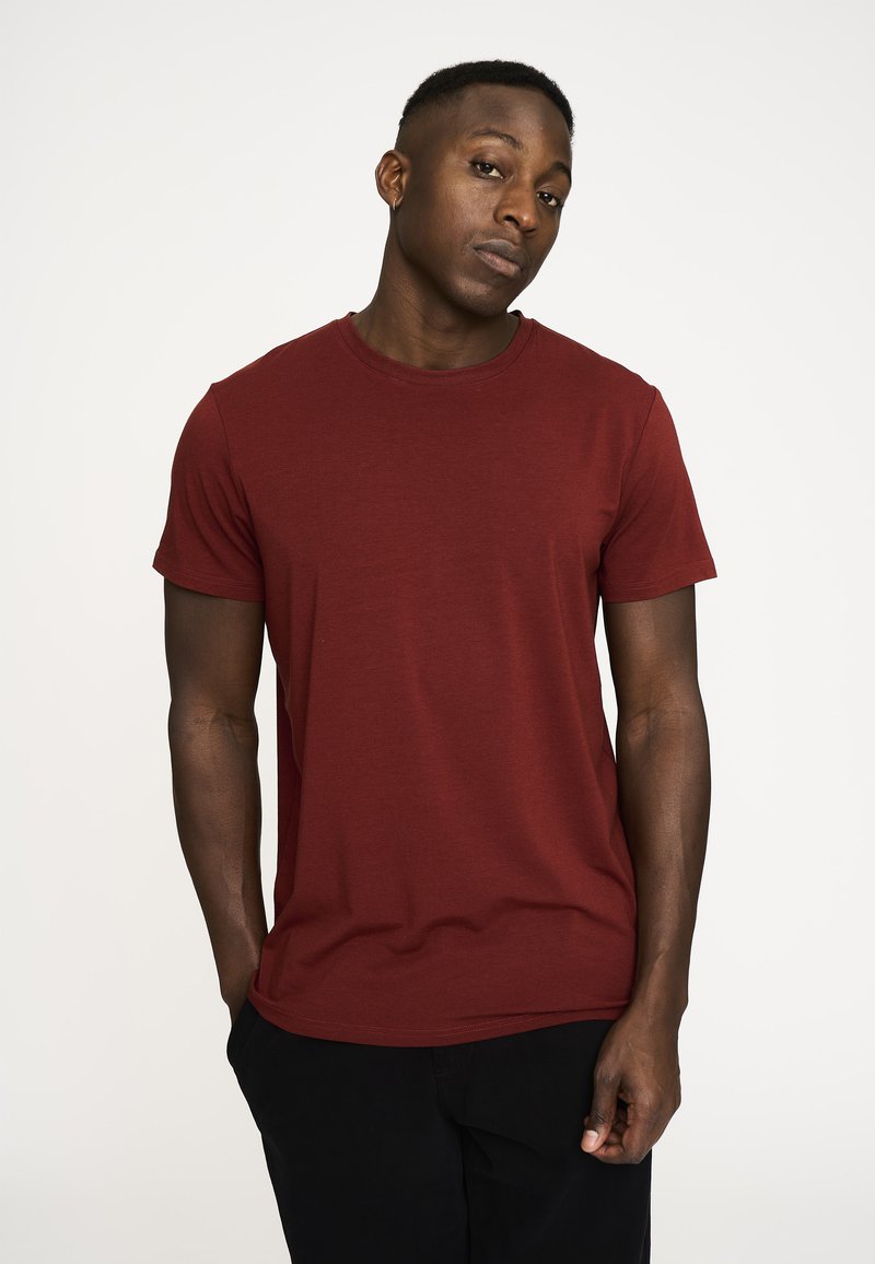 Bordeaux kortærmet t-shirt med rund hals, lavet af et blødt materiale. Båret med sorte bukser, præsenteret mod en neutral baggrund.