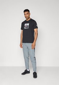 Zwarte katoenen t-shirt met een grafische print op de voorkant, gecombineerd met lichtgrijze trainingsbroek met zijstrepen en zwarte schoenen.