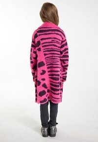 Cardigan crvene i crne boje s uzorcima nalik na krzno; dugi rukavi, otvorenog prednjeg dijela i opuštenog kroja, uparen s crnim hlačama i debelim cipelama.