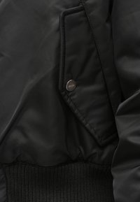 Buzunarul unei jachete negre cu clapetă fixată cu un nasture metalic gravat cu inscripția „HUGO”, având o talie tricotată cu nervuri dedesubt.