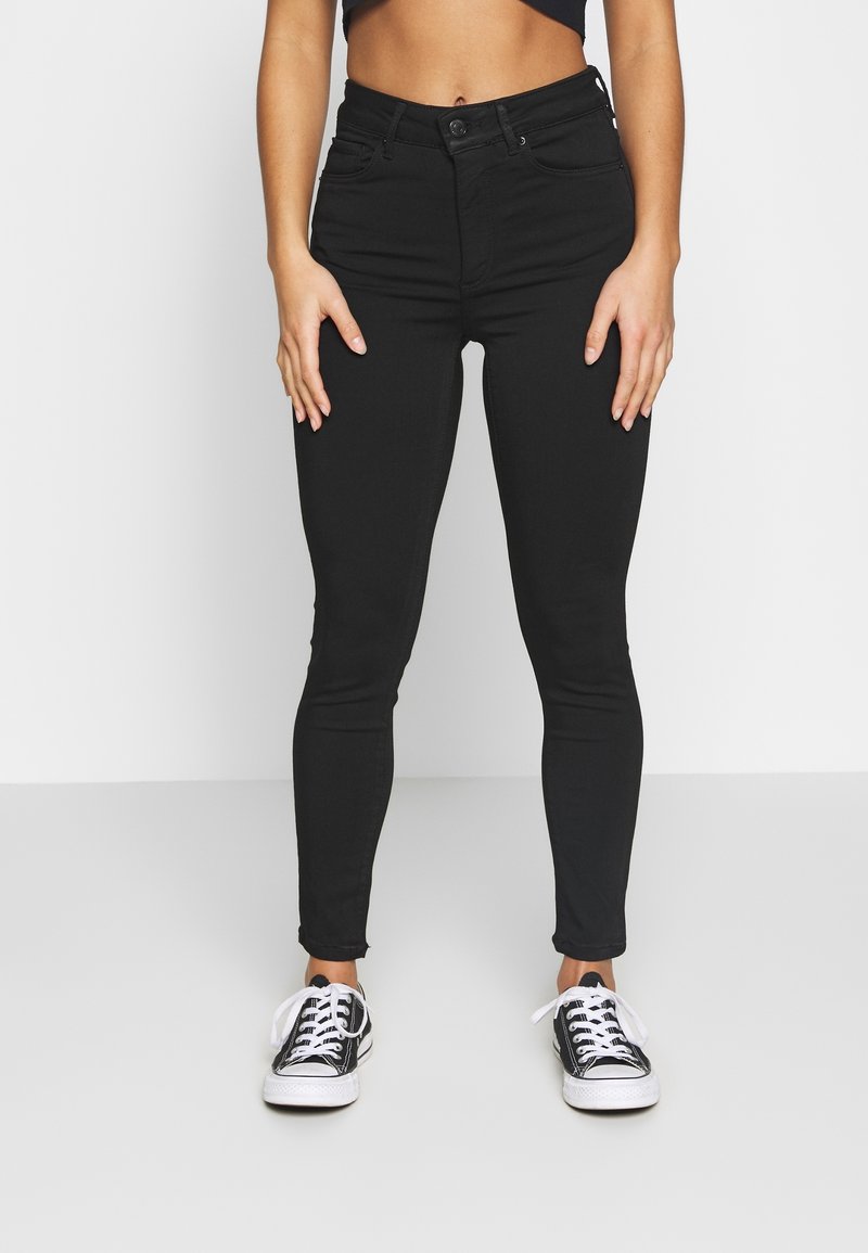 Jeans skinny negros de denim elástico, con cierre de botón y diseño de cinco bolsillos, combinados con zapatillas negras y blancas.