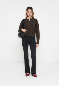 Bruine cropped cardigan met knopen, gecombineerd met zwarte flared jeans en rode schoenen. Model houdt een kleine zwarte tas vast en staat tegen een effen achtergrond.