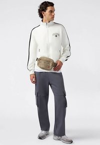 Biała kurtka na zamek z czarnymi paskami, szare spodnie typu cargo oraz beżowa torba na ramię z logo na kurtce. Szare buty sportowe.