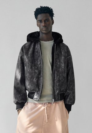 Jeune homme portant une veste à capuche noire, une chemise vert clair et un pantalon à cordon rose pâle sur un fond gris uni.