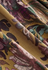 Tissu doux avec un fond vert olive et des motifs floraux abstraits en violet, sarcelle et beige, présenté avec des plis délicats et des détails de texture.