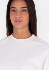 Felpa beige chiaro con scollo rotondo e maniche raglan, dotata di un piccolo logo ricamato in viola chiaro sul petto.