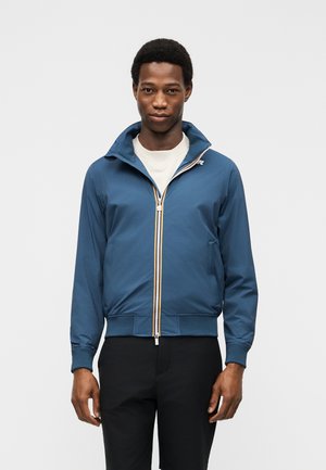 Mies, jolla on sininen vetoketjullinen takki valkoisen paidan ja mustien housujen päällä, seisoo yksinkertaisen vaaleanharmaan taustan edessä.