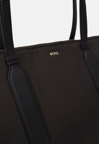 Černá taška přes rameno vyrobená z kombinace nylonu a kůže, s hladkou texturou a stříbrným detailem loga. Jednoduchý a obdélníkový design.