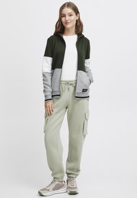 Hooded jacka i mörkgrön, ljusgrå och vit; elastiska ärmslut. Ljusgröna cargo-byxor med sidofickor. Grå sneakers med detaljer.