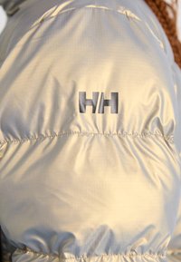 Giacca imbottita di colore chiaro con una texture liscia, un discreto motivo a quadri e un logo scuro ben in vista sulla manica.