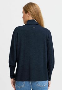 Temno modri pulover z dolgimi rokavi in visokim ovratnikom, z ribanim detajlom na manšetah in diskretnim logotipom na hrbtni strani. Mehek, teksturiran material.