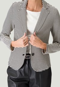 Blazer rayé en noir et blanc, doté d'un col à revers cranté, d'une fermeture à un bouton et d'une coupe ajustée. Porté sur un haut blanc et un pantalon noir.