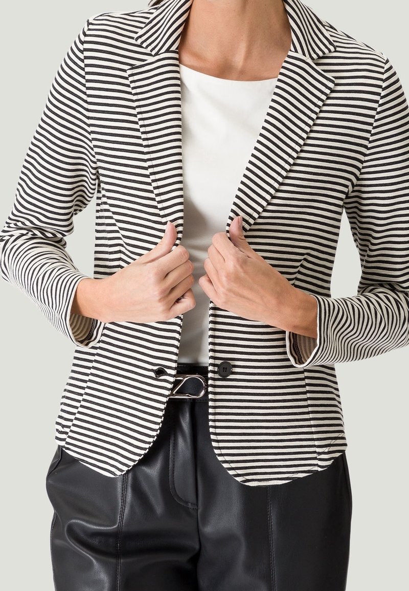 Blazer rayé en noir et blanc, doté d'un col à revers cranté, d'une fermeture à un bouton et d'une coupe ajustée. Porté sur un haut blanc et un pantalon noir.