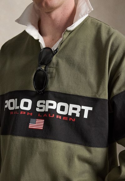 Άνδρας που φοράει πουκάμισο Polo Sport Ralph Lauren σε ελιά πράσινο και μαύρο με λευκό γιακά και μαύρα γυαλιά ηλίου κρεμασμένα στο ντεκολτέ.