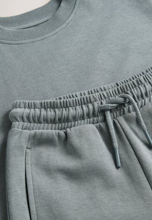 Set format din bluză sport și pantaloni sport din țesătură moale de culoare gri deschis. Guler și talie cu dungi elastice, cu șnur reglabil și buzunare laterale.