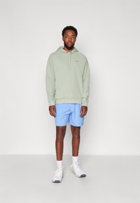 Hoodie vert clair avec une poche avant, accompagné de shorts bleus. Les baskets sont blanches avec des accents gris. Le modèle se tient devant un fond uni.