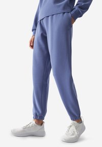 Pantalones de chándal azul claro con puños elásticos, con un acabado textil suave. Combinados con zapatillas blancas. Diseño simple y ajuste holgado.