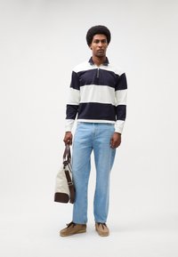 Maglia a maniche lunghe a righe blu navy e bianche con zip a metà, jeans dritti azzurri chiari e scarpe beige senza lacci, mentre tiene una borsa color crema con dettagli marroni.