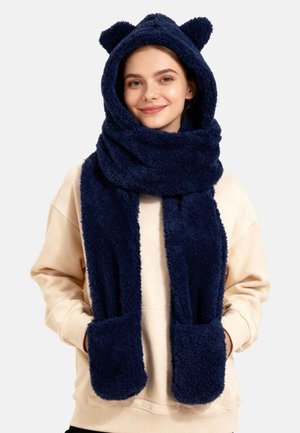 Młoda kobieta w granatowo-puszystym szaliku z kapturem z uszami misia i kieszeniami, uśmiechnięta, z rękami w kieszeniach kremowej bluzy.