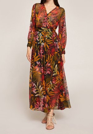 Femme portant une longue robe d'été transparente à motifs de feuilles tropicales rouges, orange, roses et vertes, associée à des sandales plates beiges.