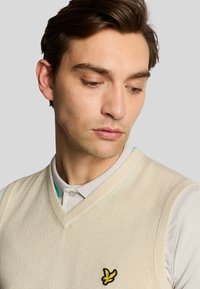 Gilet lavorato a maglia beige con scollatura a V, indossato sopra una camicia grigia con colletto. Presenta un logo di un uccello giallo e nero sul lato sinistro del petto.